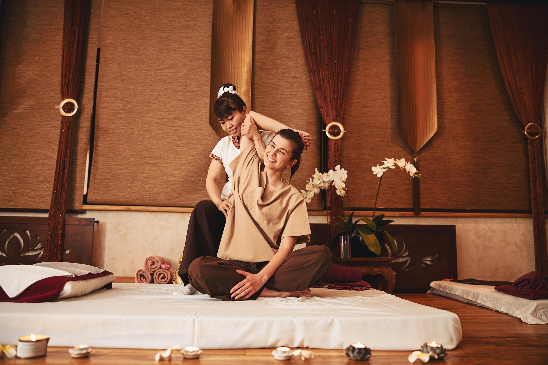 A Thai massage specialist lifts the right arm of a smiling woman while pressing her palm to the client's waist.